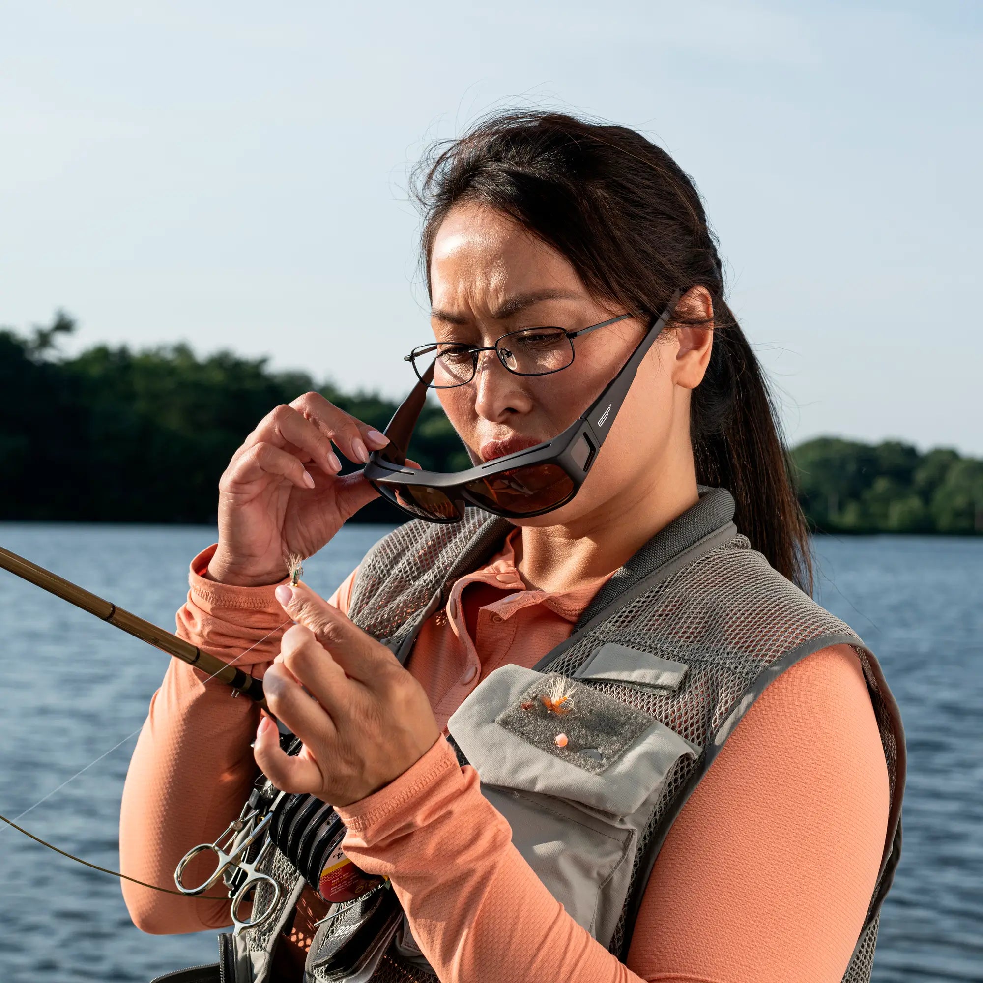 Person by water, wearing glasses and lifting flip-up shades while threading a fishing line.
